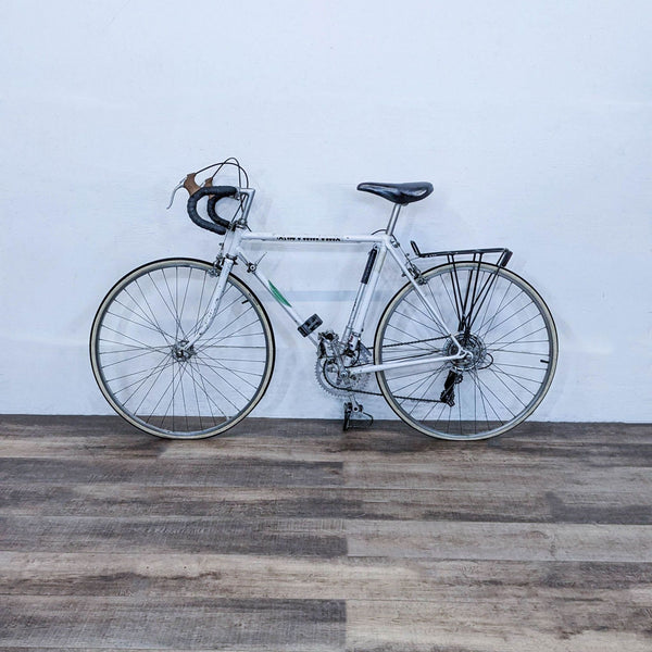 Alt text 1: A white Reperch road bicycle standing against a white wall on a wooden floor, equipped with a rear rack.