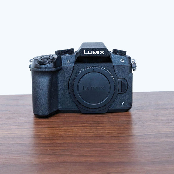 1. Panasonic LUMIX G Mirrorless Camera body on a wooden surface, front view, lens mount cap in place, no lens attached.