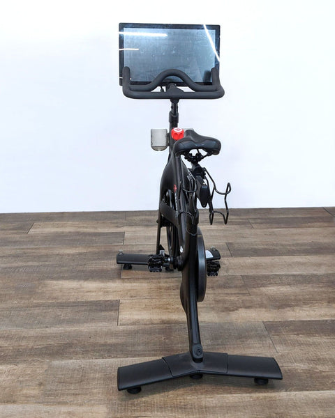 Front view of a black Peloton stationary exercise bike with a touchscreen display on a wooden floor against a white wall.