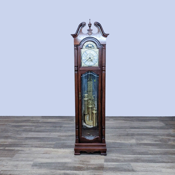 Howard Miller wooden grandfather clock with ornate face, standing on a wooden floor against a pale background.