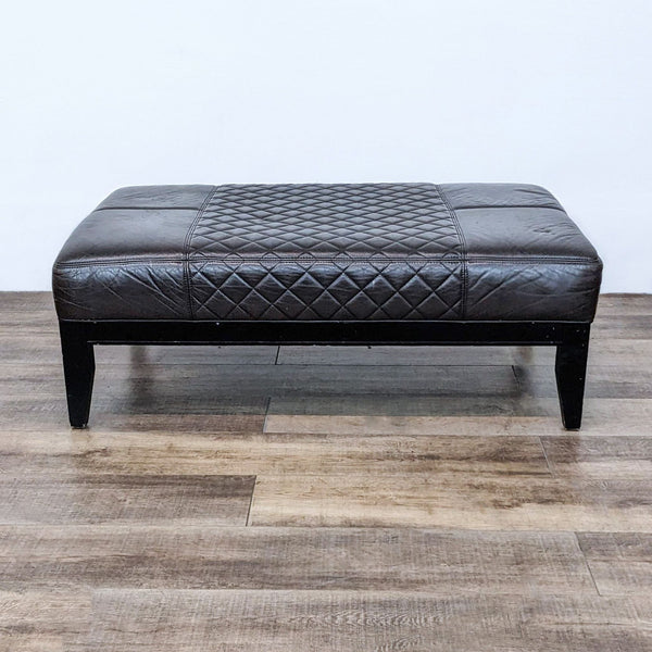 Alt text 1: Brown quilted leather ottoman with dark wood legs on a wooden floor, viewed from the side.