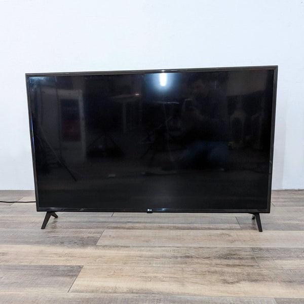 LG brand TV displayed frontally on a wooden floor against a white wall, screen off, reflecting the room.