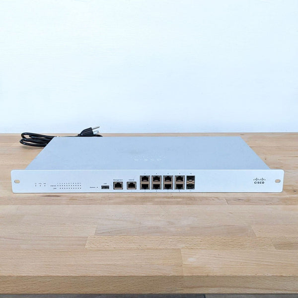 Cisco networking device on a wooden surface, front view, displaying ports and indicators. Branding visible.