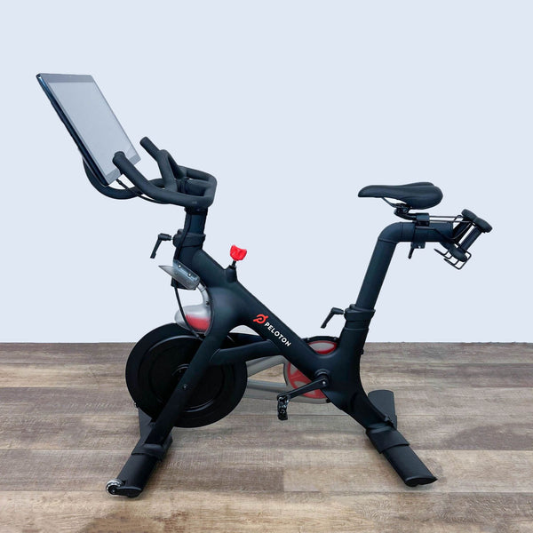 Peloton stationary bike with touchscreen monitor on a wood floor, side view.