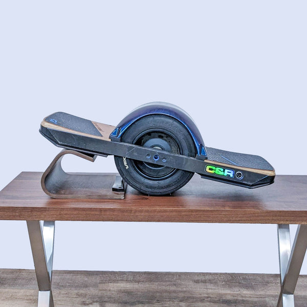 Onewheel brand gym equipment with a single large wheel centered on a wood and metal board on a table.