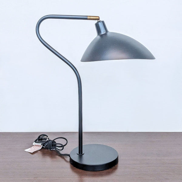 Alt text 1: A Safavieh brand desk lamp with a curved black design and a domed shade, displayed on a wooden surface.