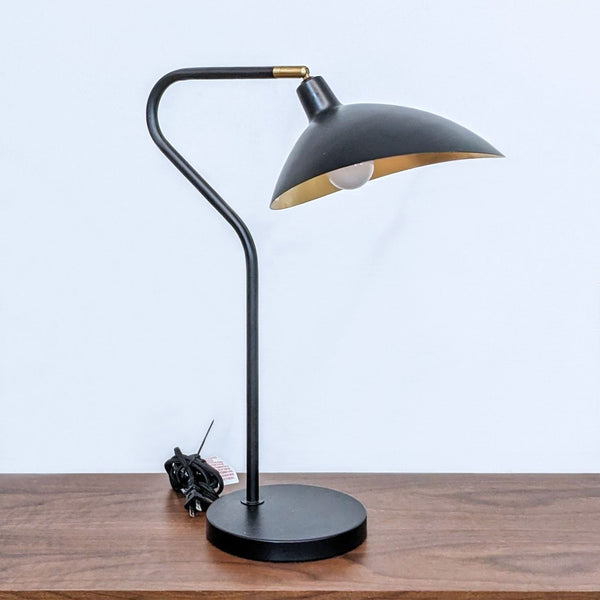 Safavieh brand desk lamp with a curved black and gold design, positioned on a wooden table against a white background.
