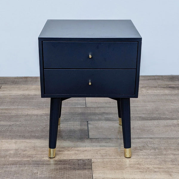 Safavieh end table with two drawers, angled legs and metallic caps on a wooden floor.