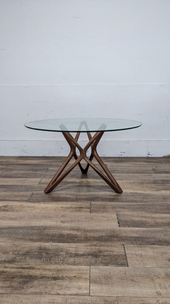 Scandinavian Design round glass tabletop dining table with a stylish wooden cross base, set against a white wall on wooden flooring.