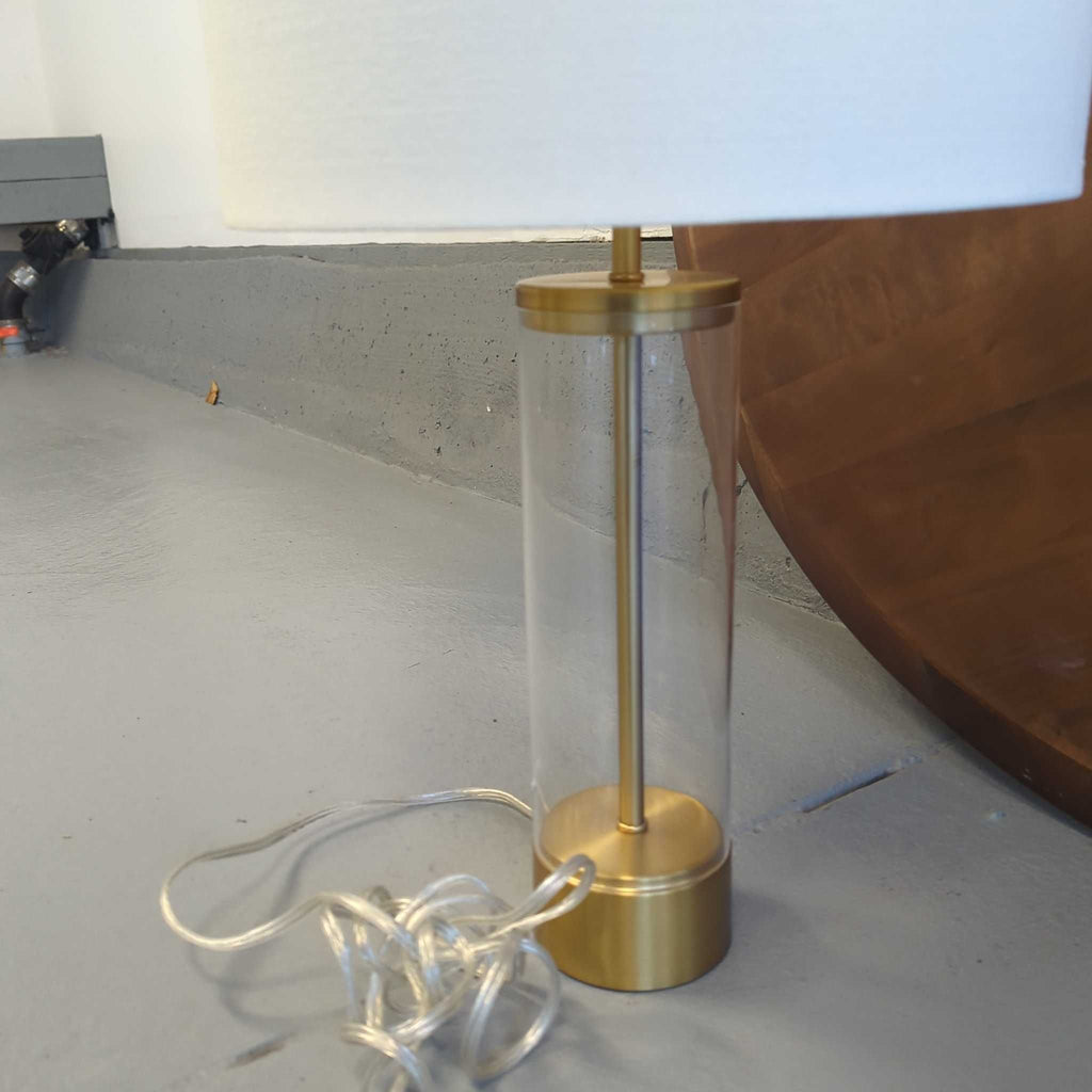 3. Elegant white-shaded table lamp featuring a clear cylindrical base and gold elements, placed next to a wall.