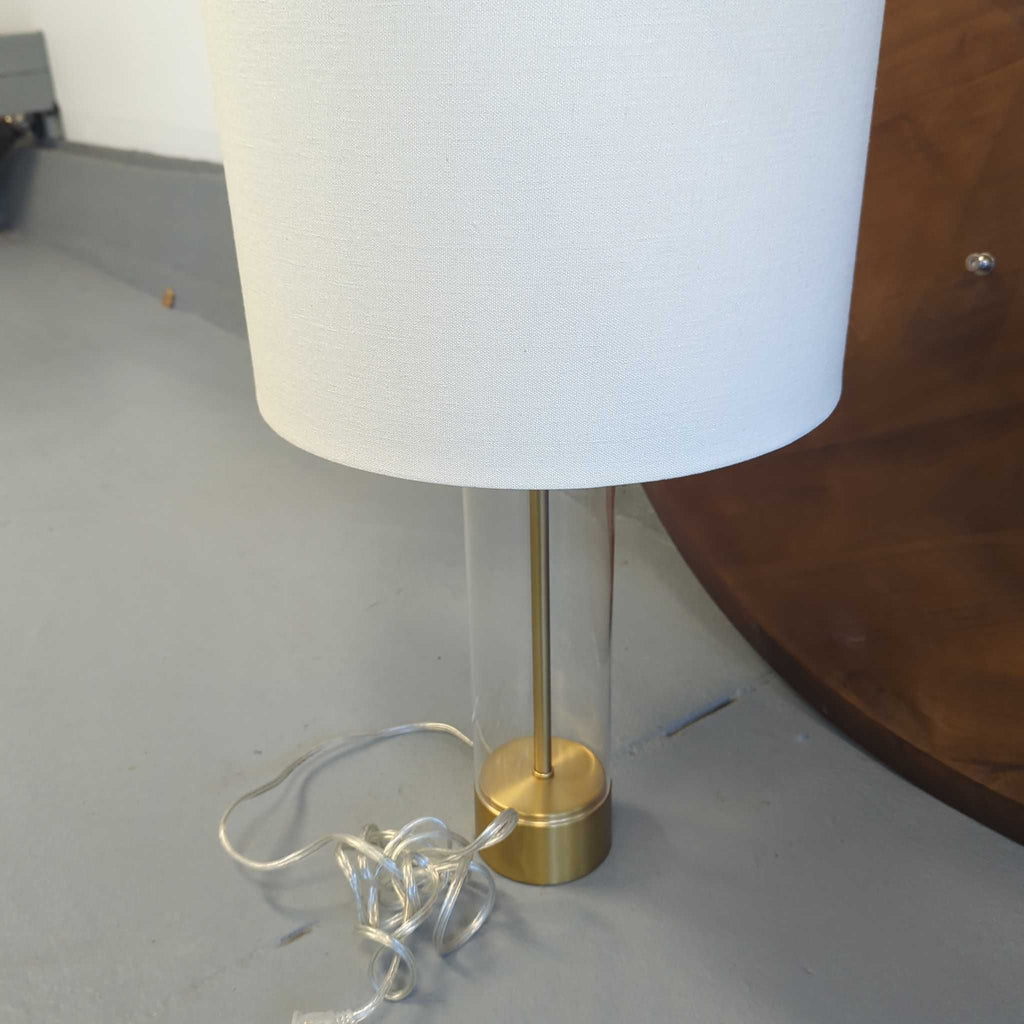 2. Close-up of a gold and glass table lamp with a white drum shade and transparent cord, for indoor lighting.