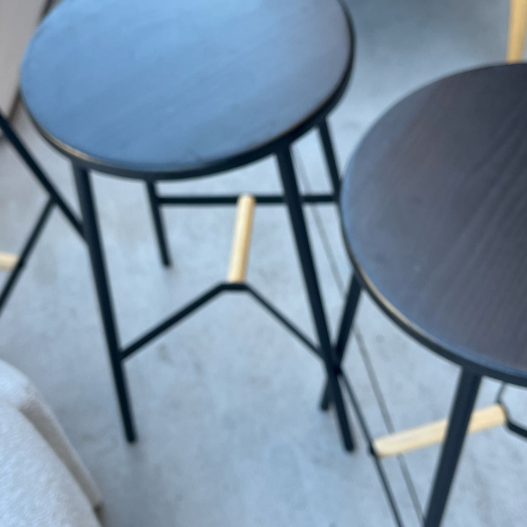 Two black round stools with metal legs and light wooden footrests on a concrete surface.  