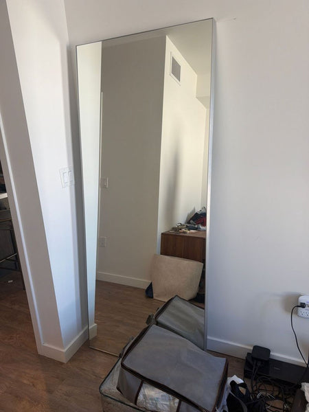 1. Tall rectangular Reperch mirror leaning against a wall, reflecting a room with wood flooring and furniture.