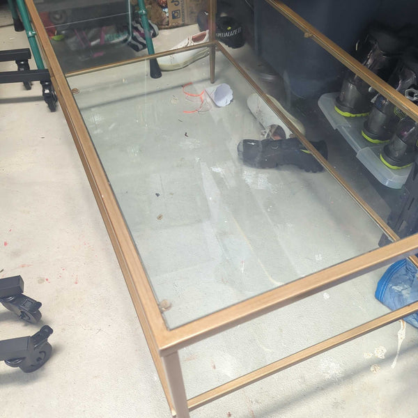 1. Rectangular coffee table with a gold metal frame and glass top, shown in a cluttered storage area.
