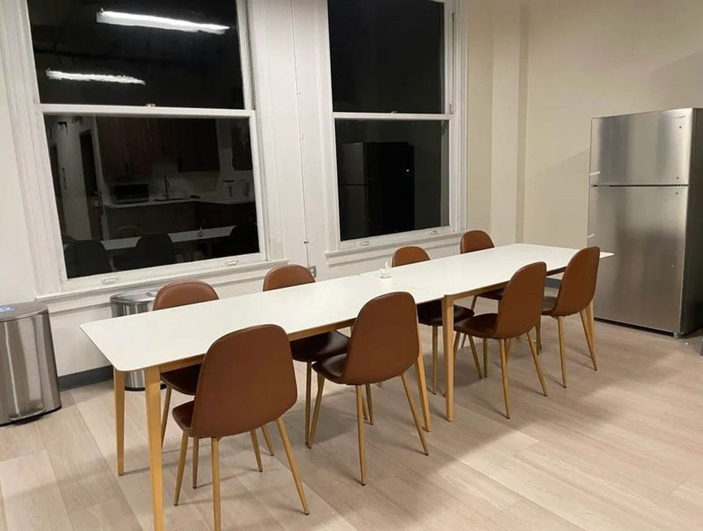 2. Long white dining table with wooden legs, paired with six brown chairs in front of large windows and a fridge.