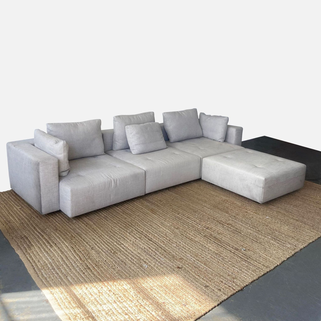 Minotti sectional with tufted design, loose back cushions, and ottoman section on a woven carpet.