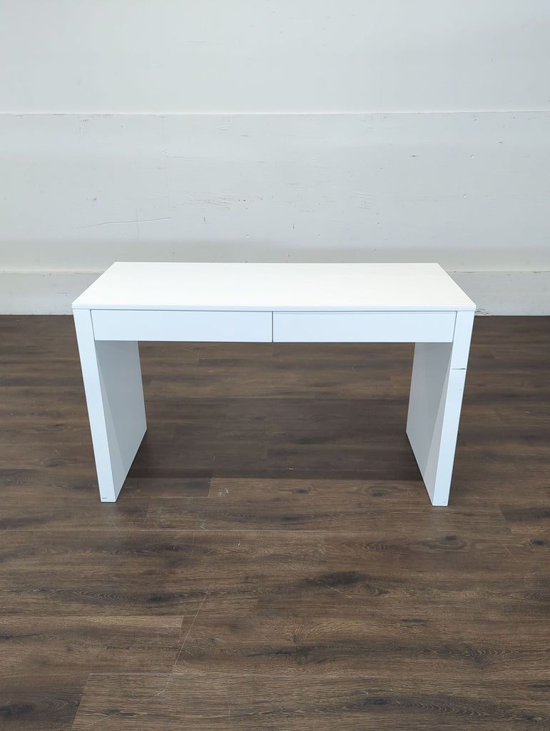 3. Sleek CB2 white desk with a simple rectangular shape and two drawers, photographed against a blank wall.
