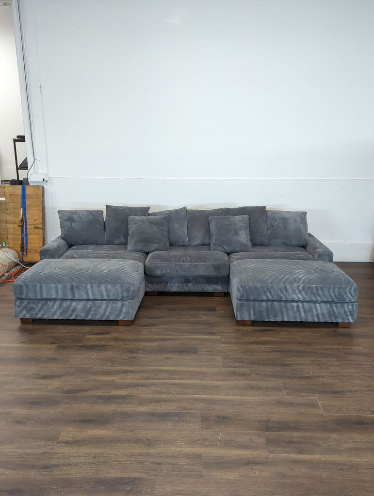 3. OAH Furniture grey 3-seat sectional sofa with ottoman, displayed in an empty room with wood flooring.