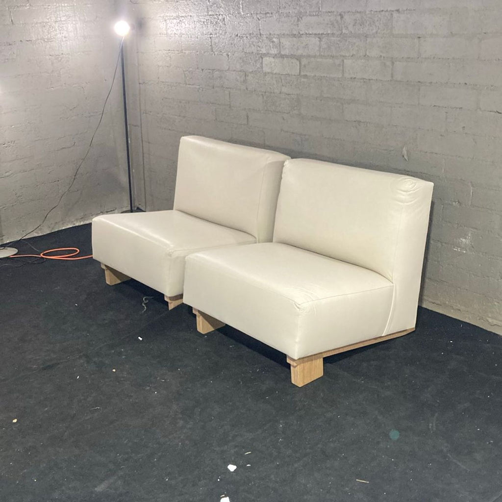 1. Two armless cream lounge chairs with wooden legs set against a gray brick wall in a minimalist room.  