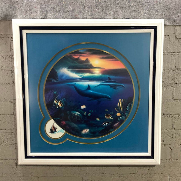 Circular framed print of dolphins and tropical fish beneath a sunset, with gold border and glossy white frame.