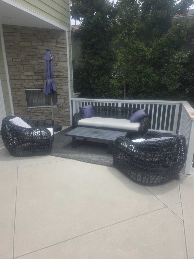 Patio furniture set featuring a cushioned wicker sofa, two chairs, coffee table, and accent pillows.