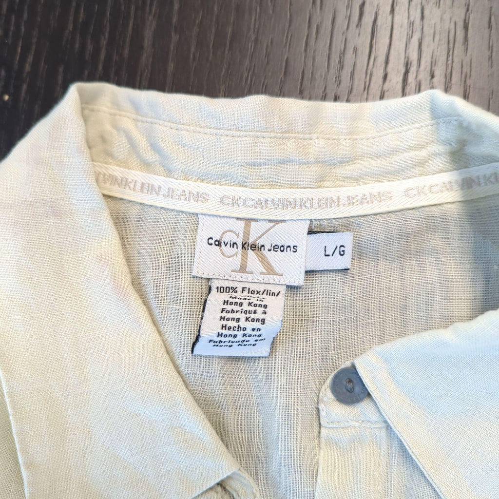2. Close-up of Calvin Klein Jeans tag and care label on a light beige women's shirt, size large.