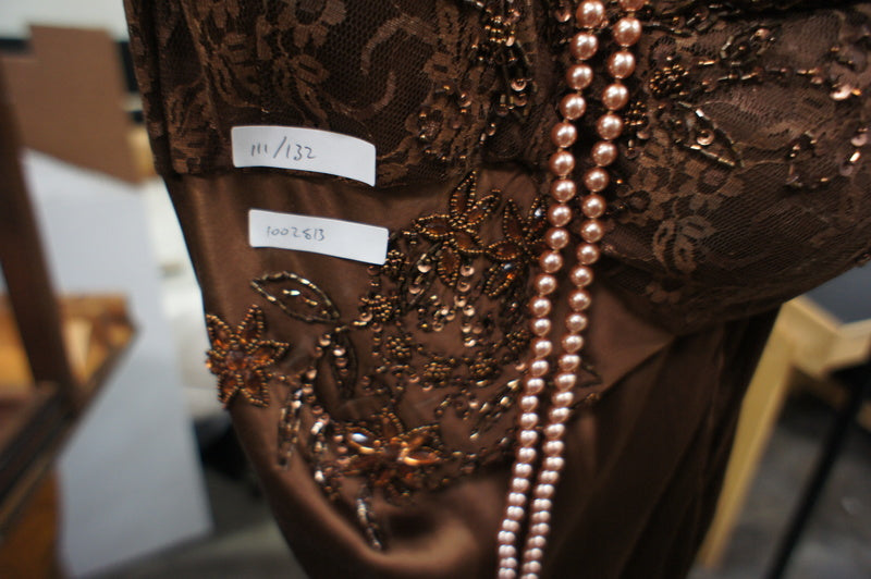 2. Close-up of brown lacy Bicici dress showing floral embroidery and long pearl necklace detail.