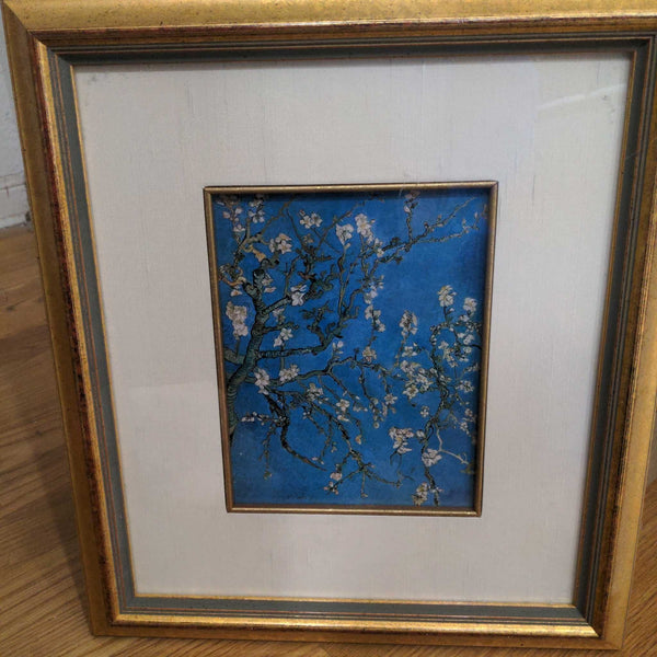 Framed art print of blossoming branches against a blue background with a gold frame, seen up close.  