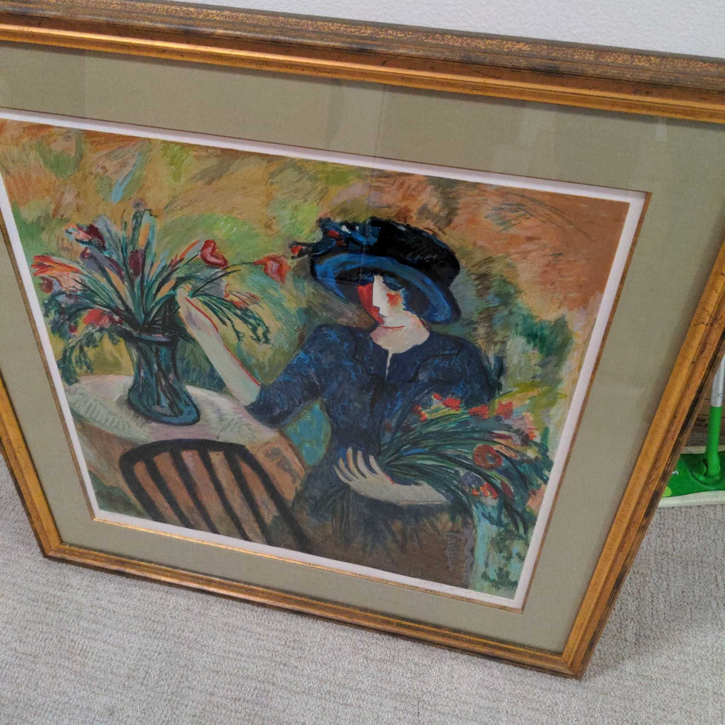 1. Framed painting of a woman in a blue dress and hat arranging flowers at a table, in an impressionist style.
