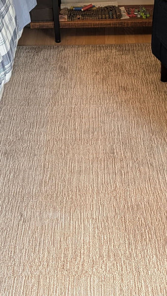 1. Beige rug with a subtle linear pattern, placed beside a bed and near a shelf in a bedroom.