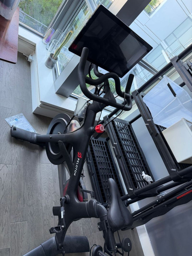 1. Peleton stationary exercise bike with touchscreen monitor, positioned indoors near large windows.