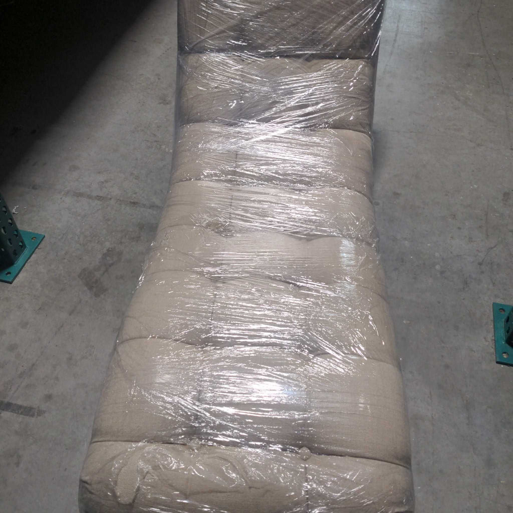 Beige tufted lounge chair wrapped in protective plastic, seen from above on a warehouse floor.  