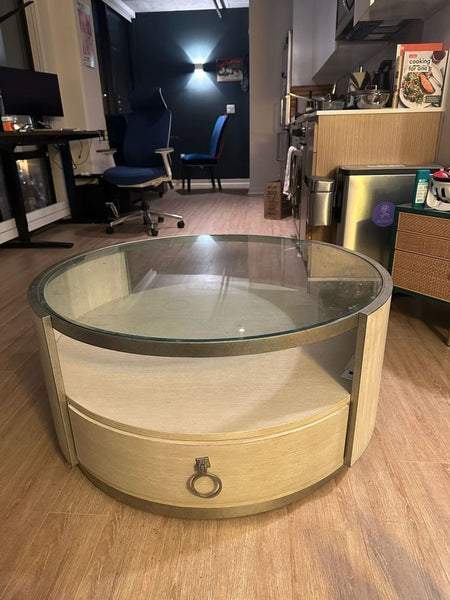 1. Round coffee table with glass top, light wood finish, lower shelf, and drawer with metal ring handle in room setting.  