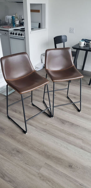 1. Pair of Reperch brown leather bar stools with black metal legs on wood floor, shown in a modern apartment setting.  