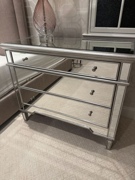 Mirrored dresser with three drawers, metallic trim, and crystal knobs, placed in a modern bedroom setting.