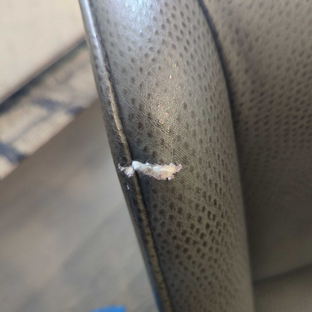3. Detailed view of a tear with exposed stuffing on the edge of the stool’s gray upholstered armrest.