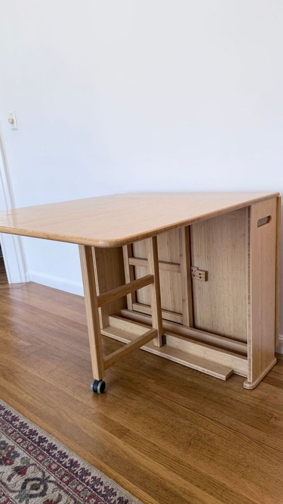 Foldable Greenington dining table extended for use, with light bamboo finish and wheels, against a white wall.  