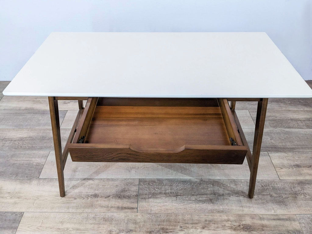 Mid-century desk with a white top and wood drawer, open on a wooden floor.