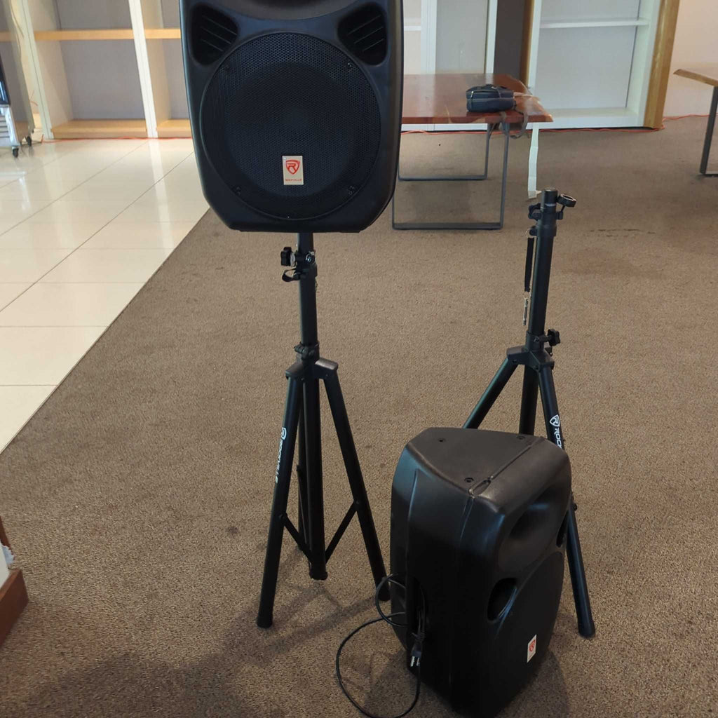2. Two Rockville speakers with tripod stands, one standing and one lying down, in a room with carpet flooring.