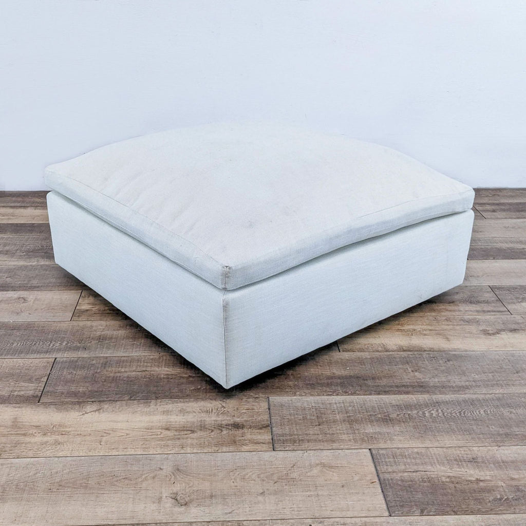 White fabric ottoman with plush cushion, square shape on wooden floor.