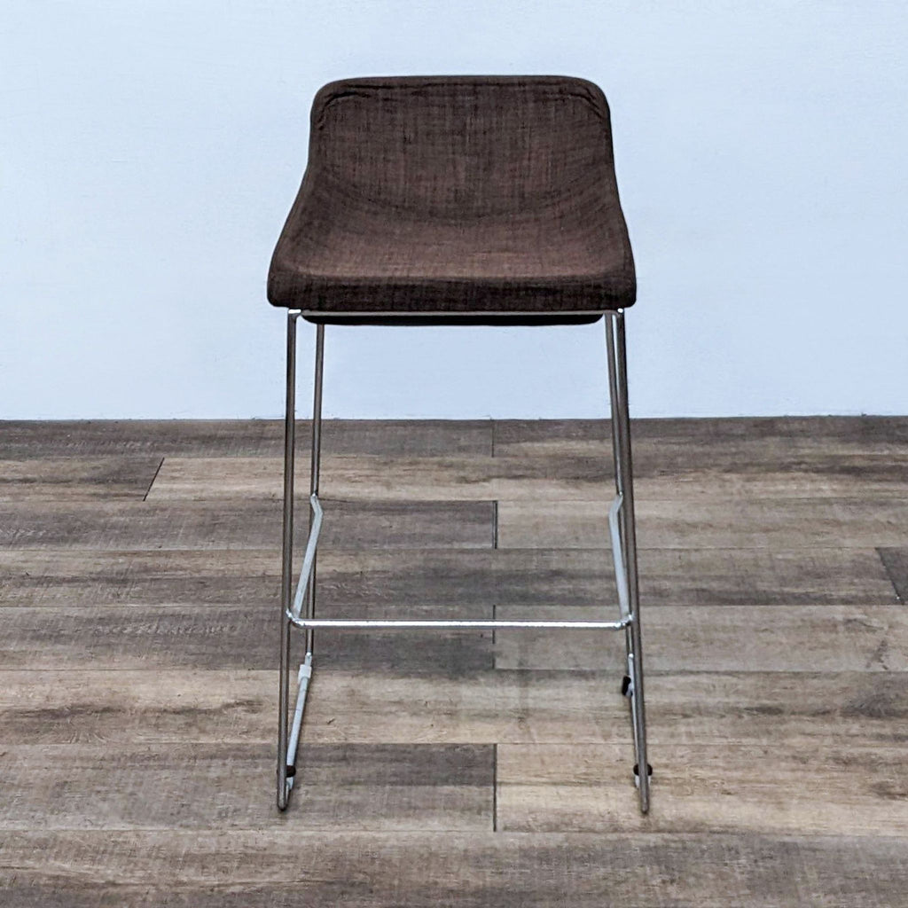 Brown fabric stool with padded cushion and chrome frame, set on wooden floor.