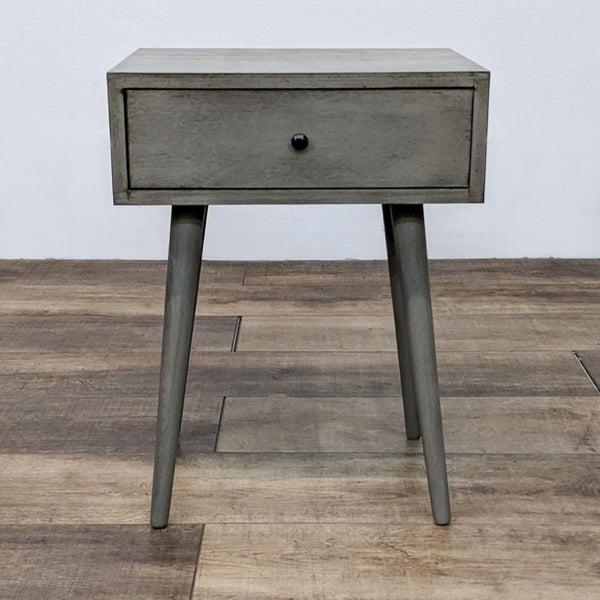 Simple grey end table with a drawer and splayed legs on wooden floor.