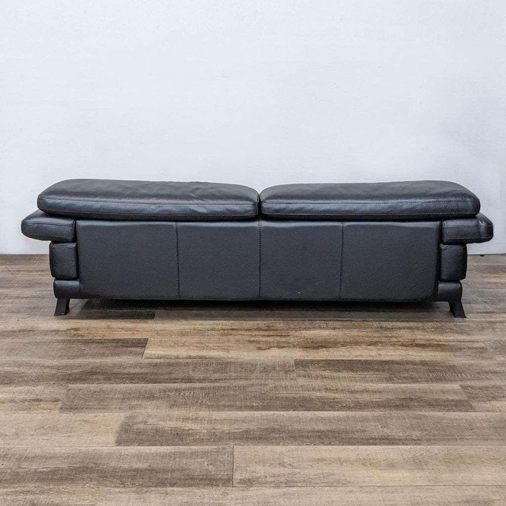 Rear view of low profile black leather loveseat on wooden floor.