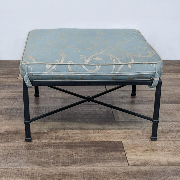 Blue cushioned ottoman with ornate pattern on a metal frame, placed on wooden flooring.
