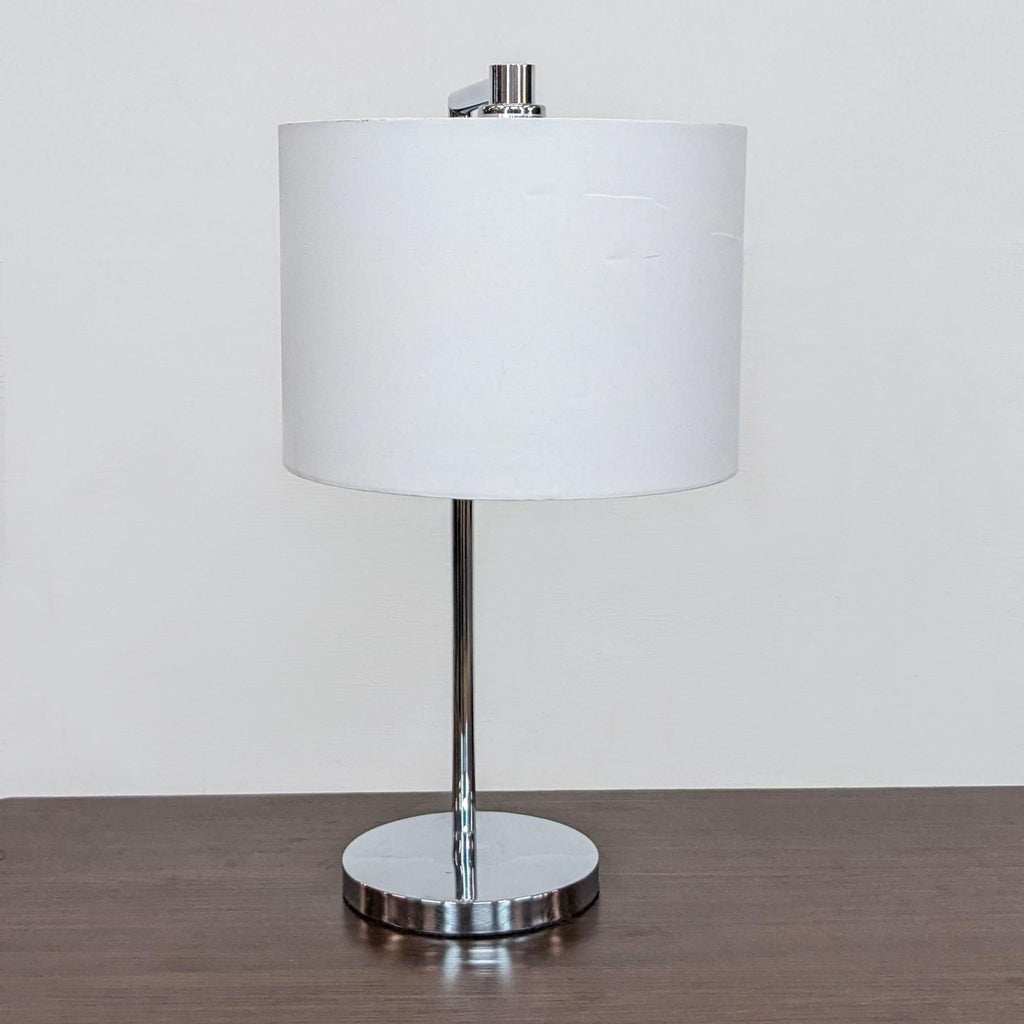 Modern table lamp with a white drum shade and chrome details by Zuo Modern on a wooden table.