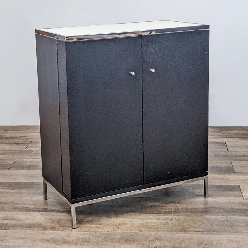 Black cabinet with metal legs and white top, closed doors. Lacks interior shelving.