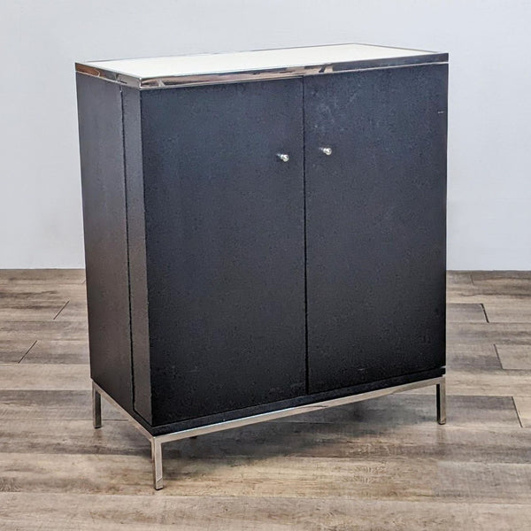 Black cabinet with metal legs and white top, closed doors. Lacks interior shelving.
