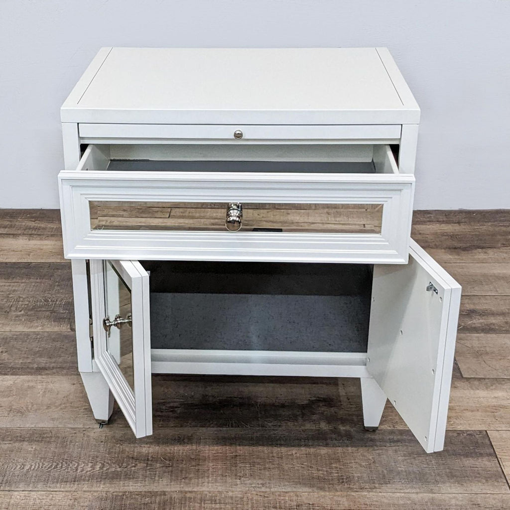 End table with open drawer and doors, featuring mirrored and moulded fronts, on a wooden floor.
