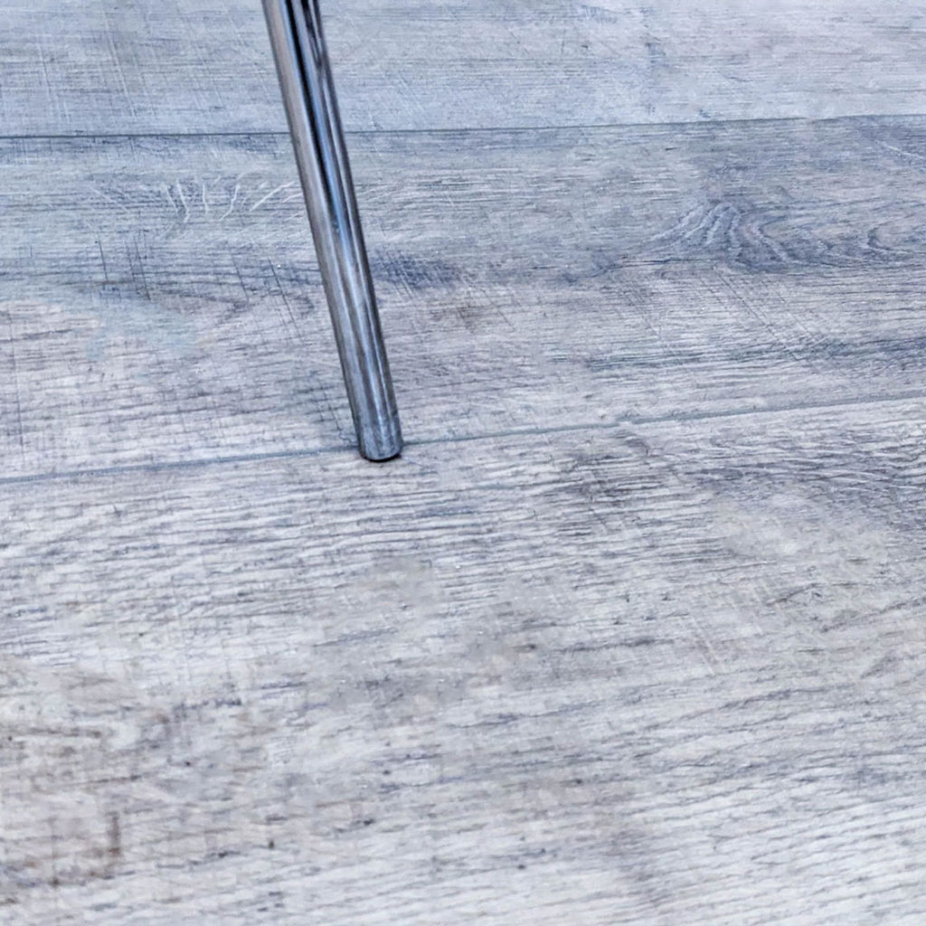 Close-up of the Luther chair's chromed leg with rubber feet on wooden flooring, from Strick & Bolton.