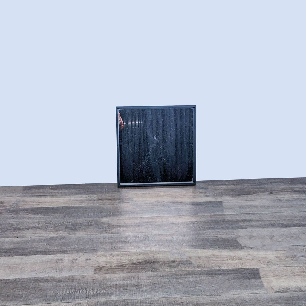 Square wall mirror with a black frame on a wooden floor.